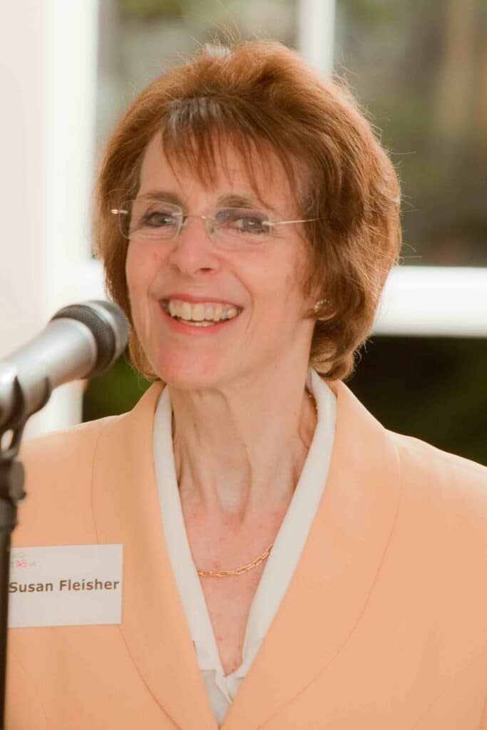 Image of a woman with short brown hair and wire frame eyeglasses smiling at a microphone. She's wearing a peach coloured suit jacked and a white blouse. She has a name tag that says Susan Fleisher
